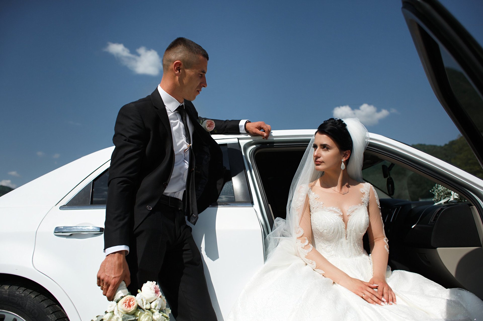 Man stands next to bride outside white car as she holds bouquet of flowers. They are in an outdoor setting under blue sky. The couple is ready for the wedding.