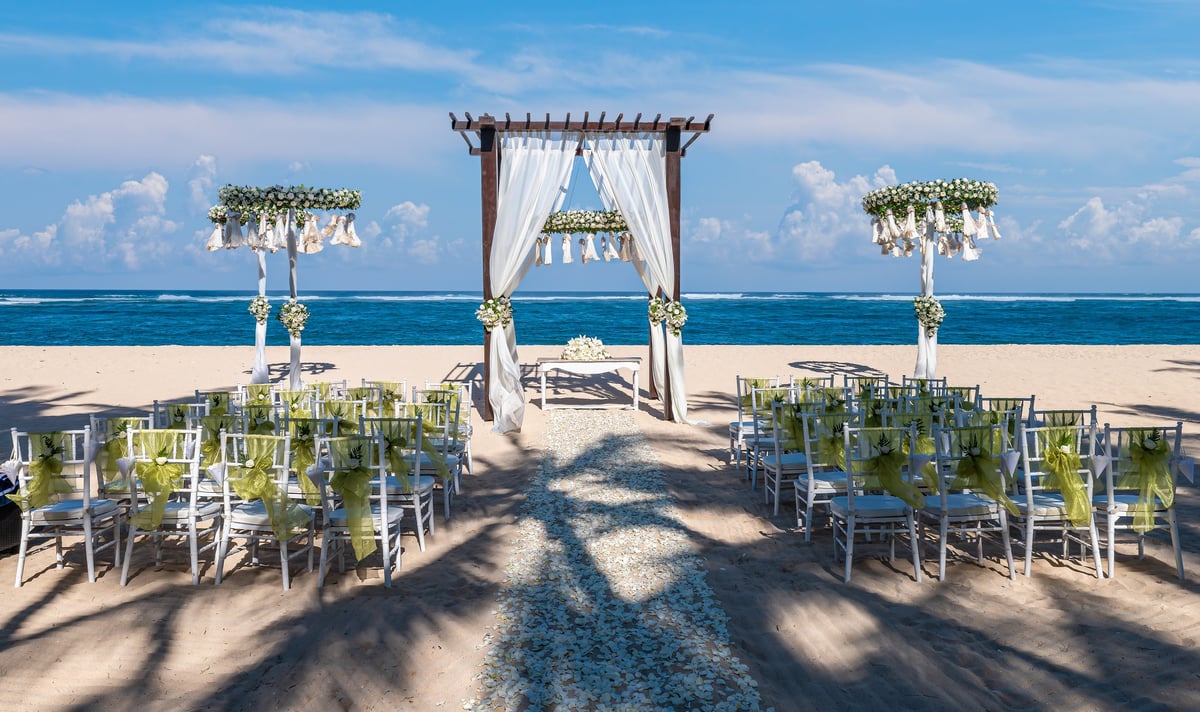 Elegant beach wedding ceremony setup on a sandy tropical shore with white floral decorations, chairs, and a romantic ocean backdrop.