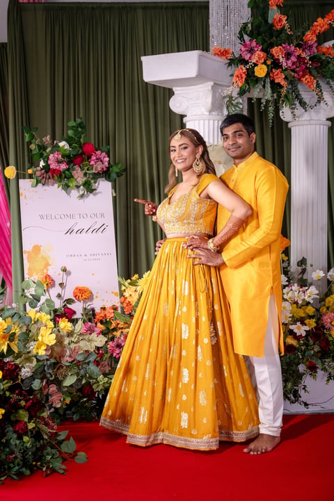Couple in matching yellow traditional Indian attire posing on red carpet with elaborate floral backdrop and welcome sign