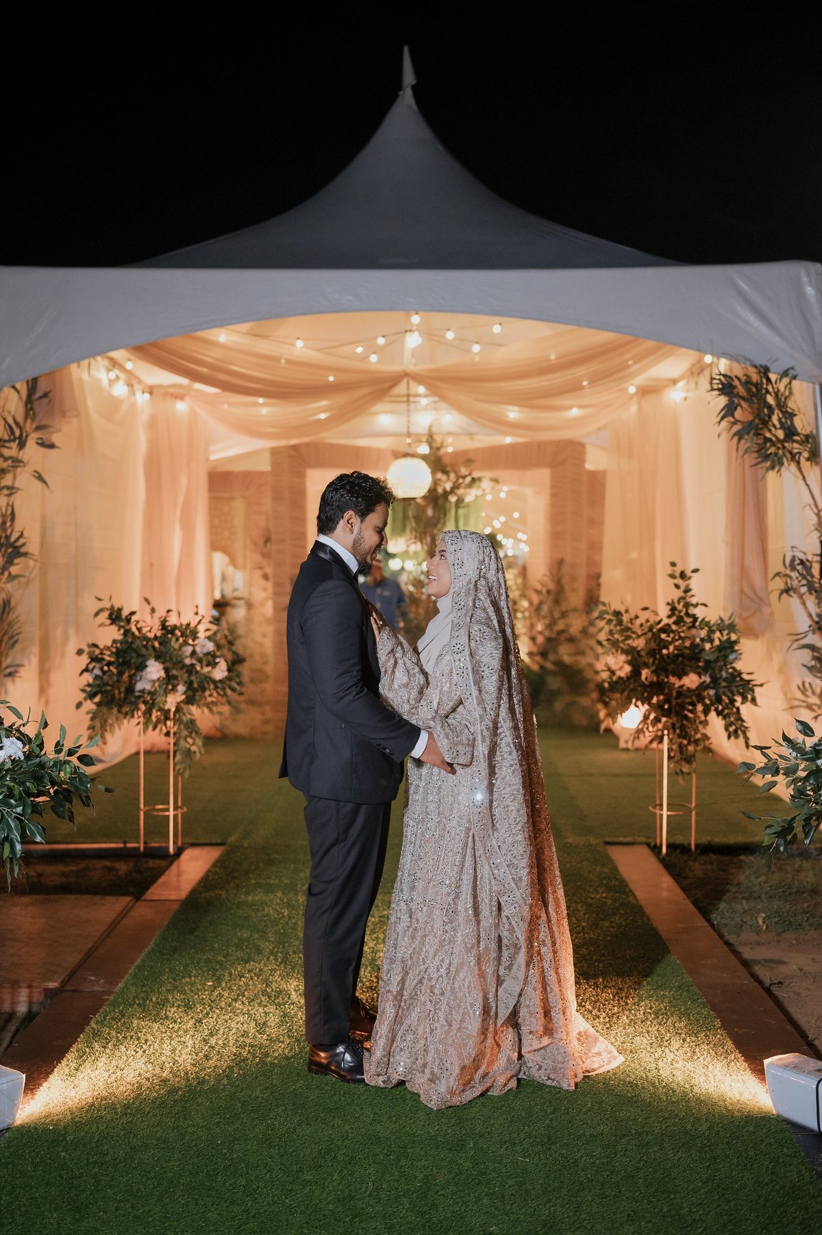 Groom in dark suit and bride in ornate shawl holding hands under elegant tent with string lights, flowers, and decorated entrance