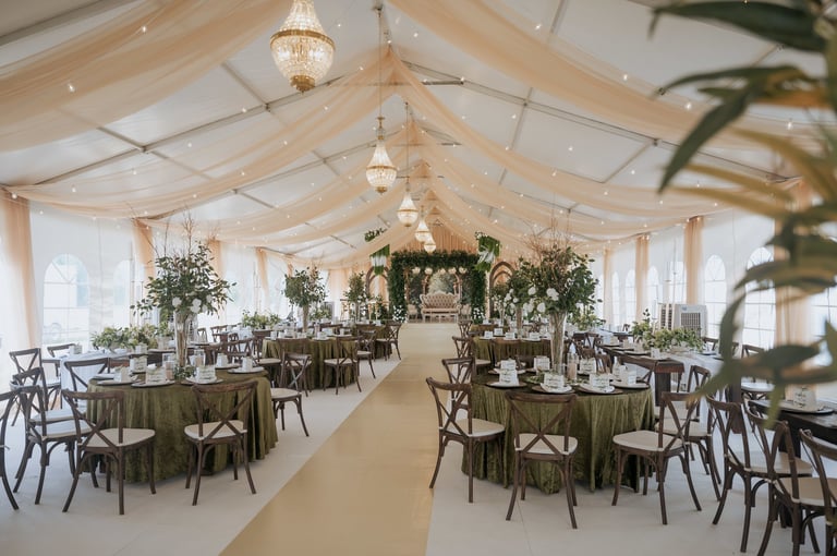 Elegant white tent wedding reception with sage green table linens, wooden chairs, hanging floral arrangements, and crystal chandeliers