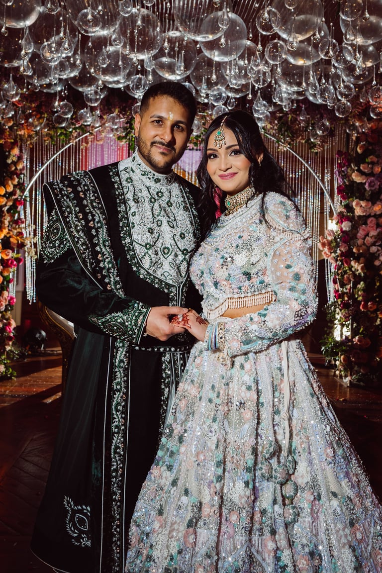 A couple in ornate traditional Indian wedding attire holding hands under crystal chandeliers and floral decorations
