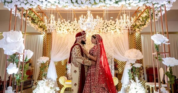 Bride and groom in traditional Indian wedding attire standing under an ornate gold and floral decorated wedding mandap