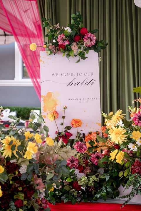 Welcome sign for a wedding event decorated with colorful flowers including pink, yellow, and red blooms, with green curtains in the background