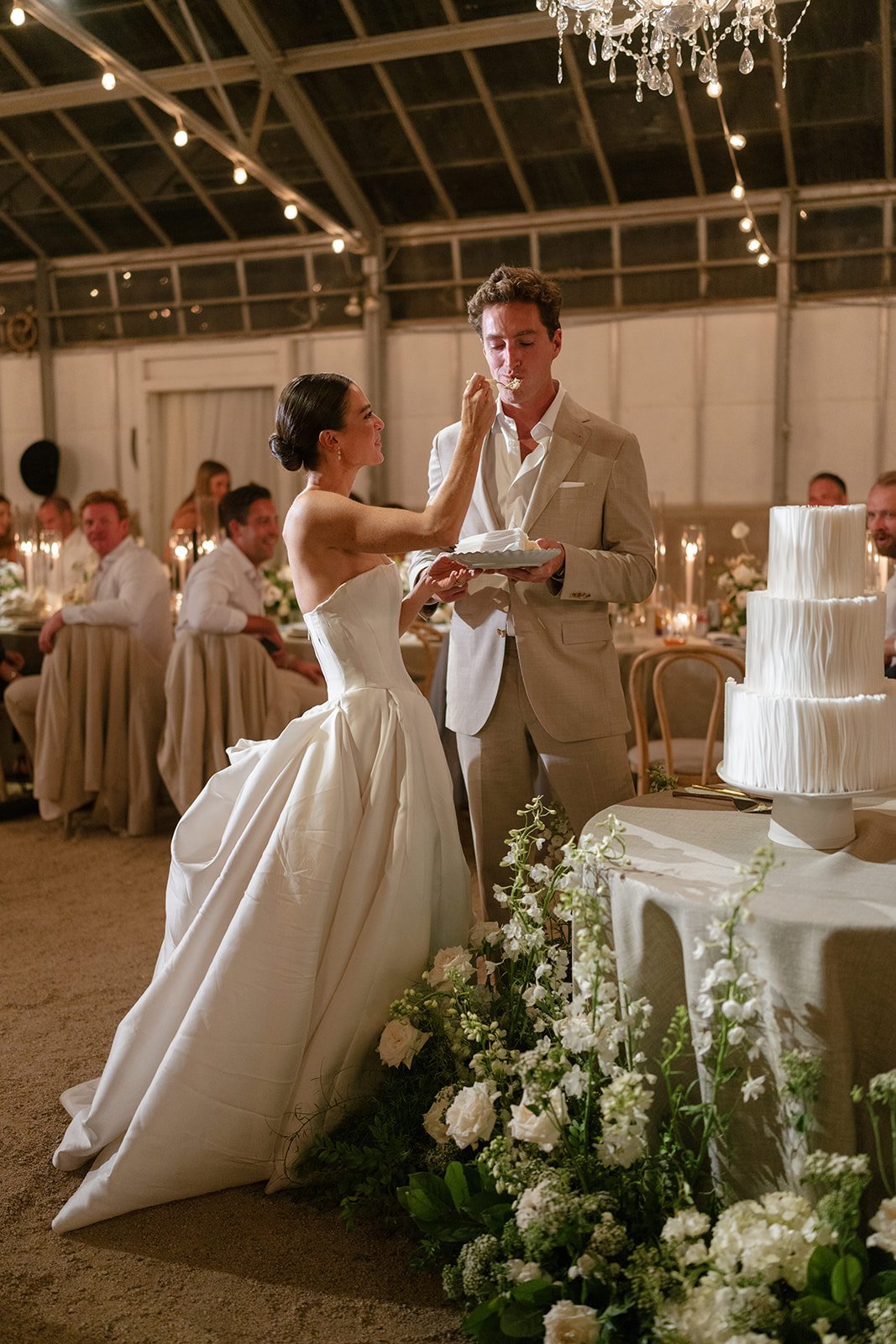 Bride and groom feeding each other cake at wedding reception in elegant greenhouse venue with string lights and white floral decorations