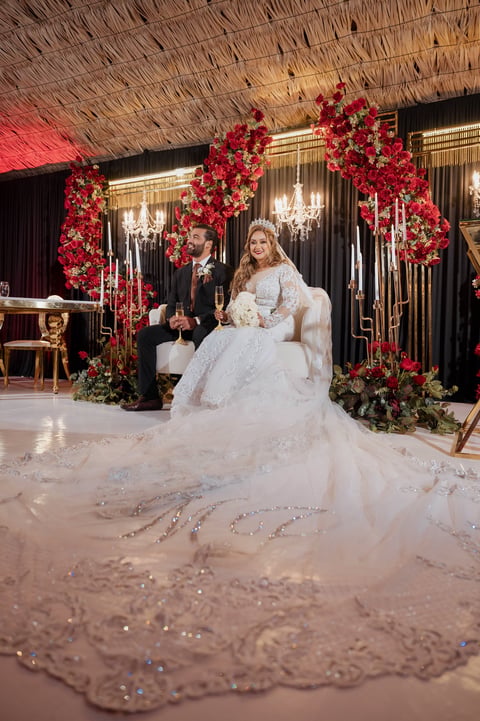 Bride in elaborate white gown with beaded train seated near groom under red floral arch at elegant wedding reception