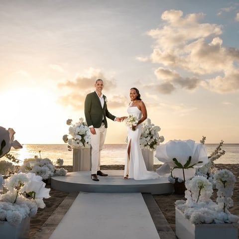 Couple exchanging vows on beach platform during sunset wedding ceremony with white floral decorations