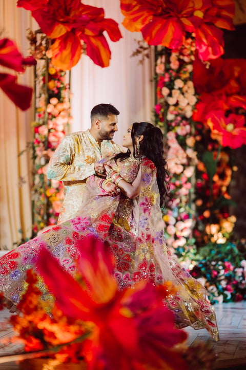 A couple in traditional wedding attire stands together surrounded by vibrant red and orange floral decorations and arrangements.