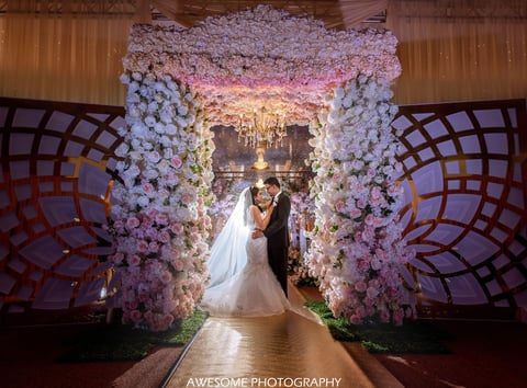Bride and groom standing under an ornate floral archway with white and pink flowers and golden chandelier in ballroom