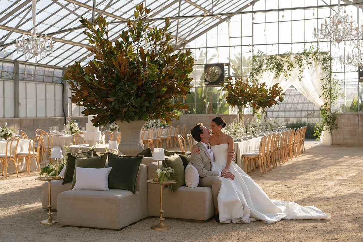 Bride and groom sitting on beige lounge seating in modern greenhouse wedding venue with large floral arrangement and rows of chairs
