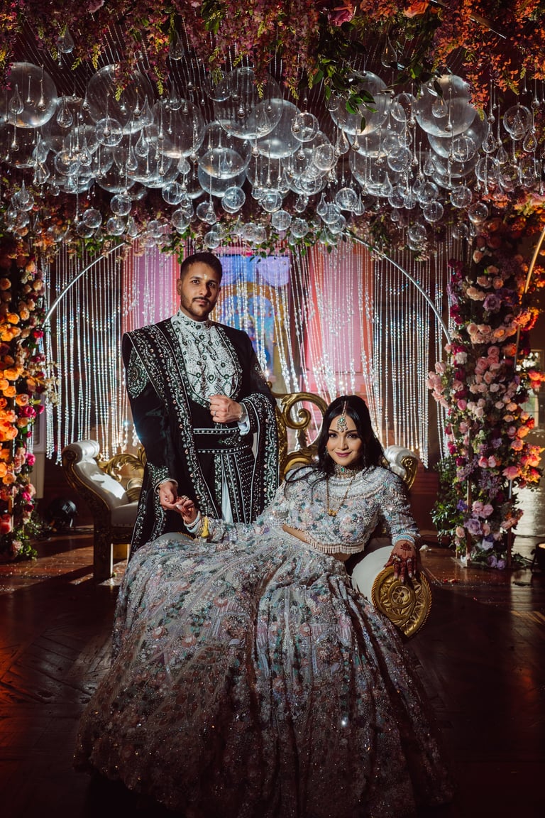 A couple in ornate traditional wedding attire posing under an elaborate floral archway with hanging chandeliers and pink lighting