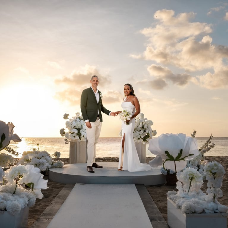 Bride and groom exchange vows on a raised platform during beachside wedding ceremony at sunset with white floral arrangements