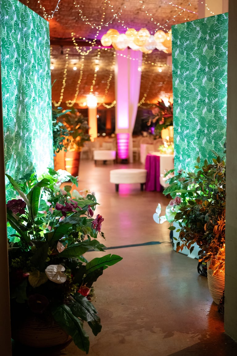 Illuminated event venue entrance with green patterned walls, pink and gold lighting, decorative plants, and a lit archway leading into a festive space