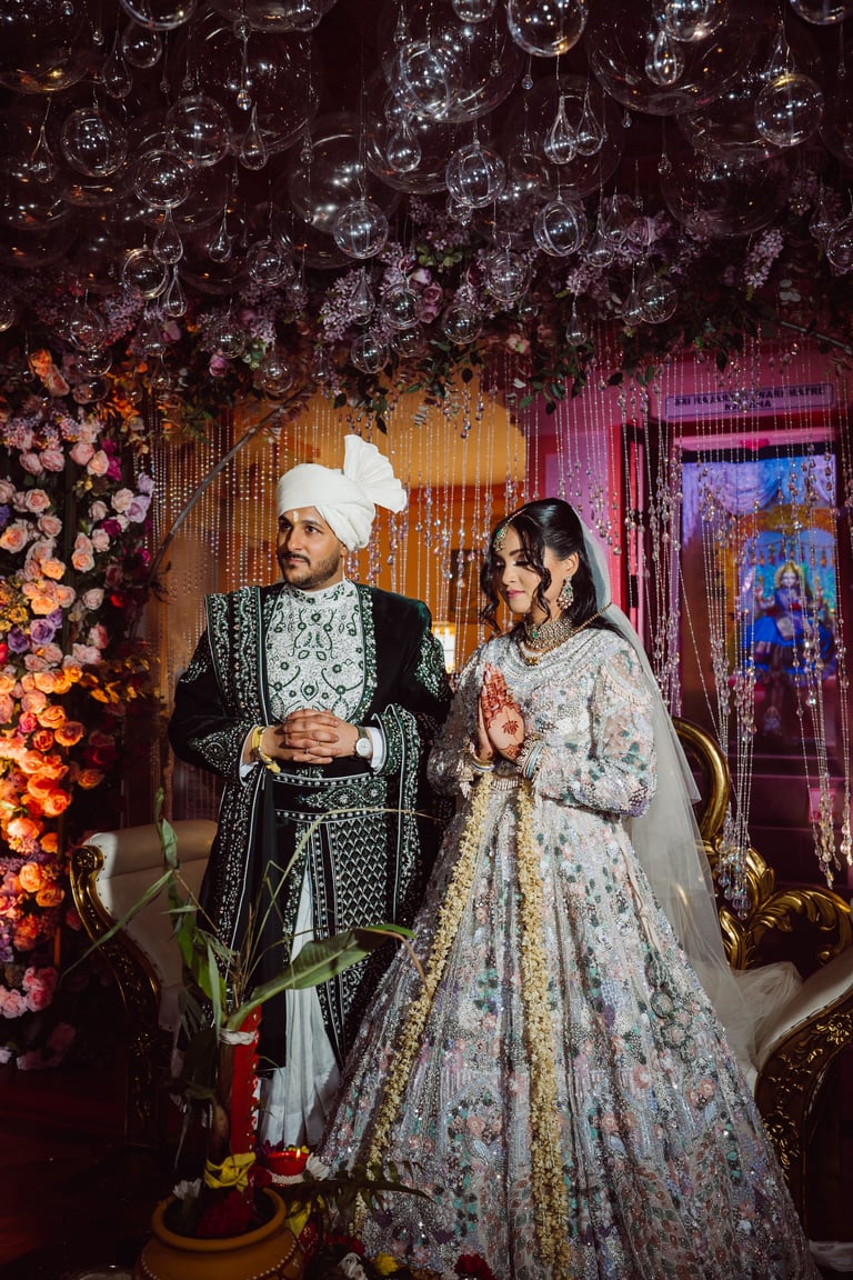 Couple in traditional Indian wedding attire at ornate ceremony with hanging crystal decor, floral arrangements, and colorful lighting