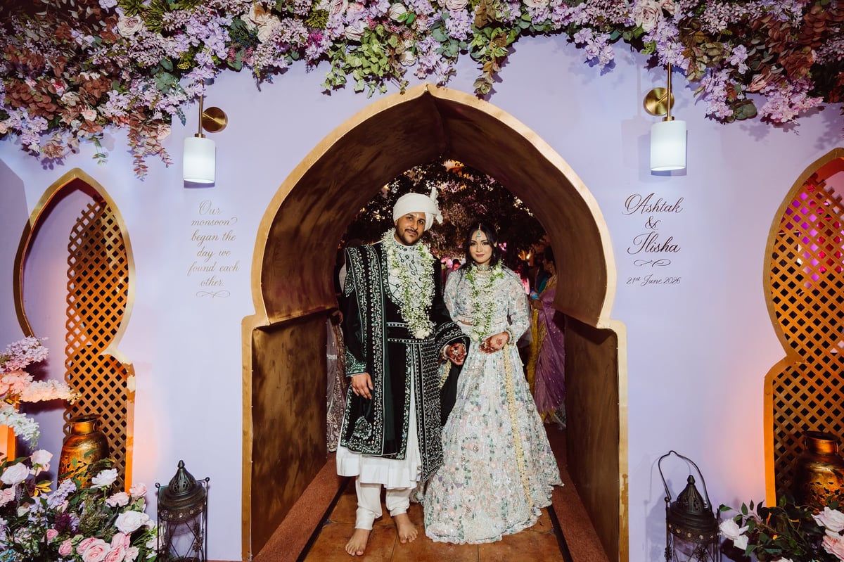 Bride and groom in traditional Indian wedding attire posing in a ornate archway decorated with pink flowers and hanging lanterns