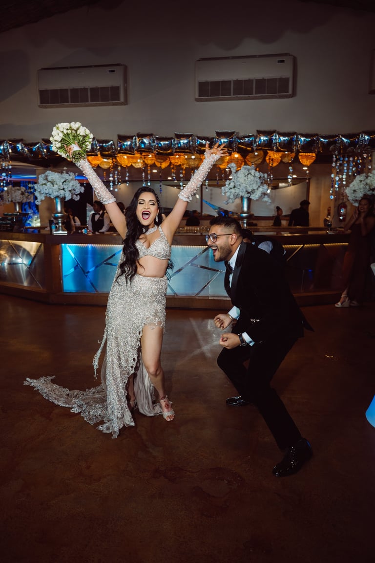 Bride in sparkly wedding dress with arms raised dancing with groom in tuxedo on decorated ballroom dance floor