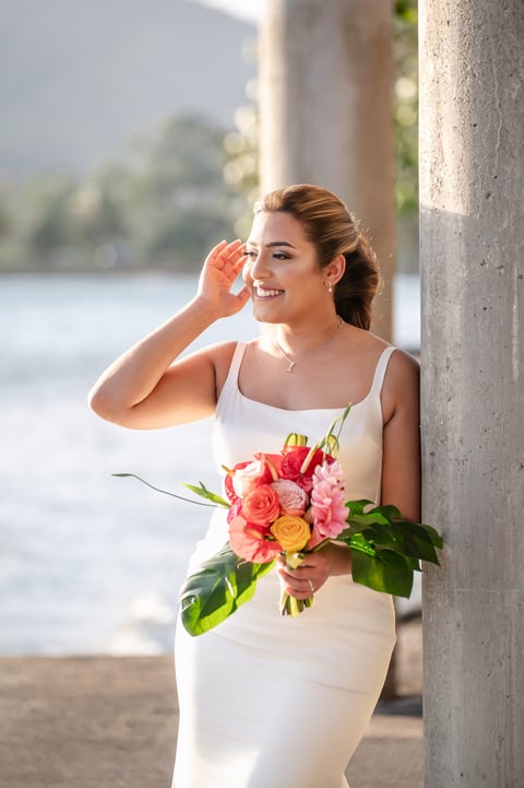 Woman in white dress holding colorful bouquet of roses, smiling by concrete pillar with waterfront view