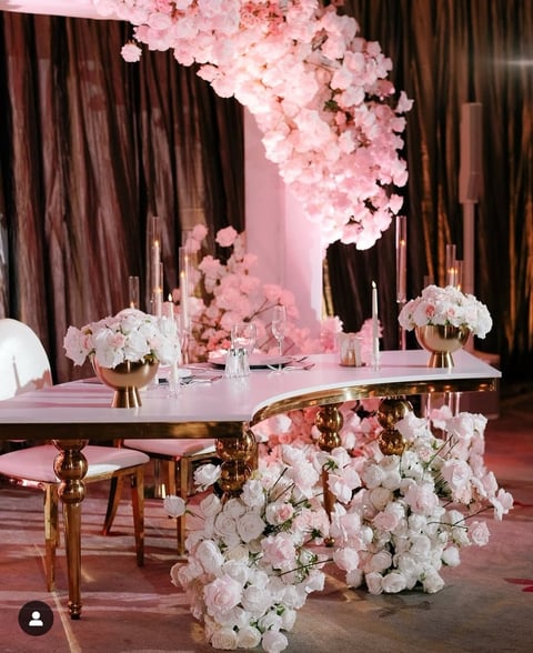 Elegant wedding reception setup with pink and white floral arrangements, luxurious gold-accented furniture, and draped brown curtains in a sophisticated venue.