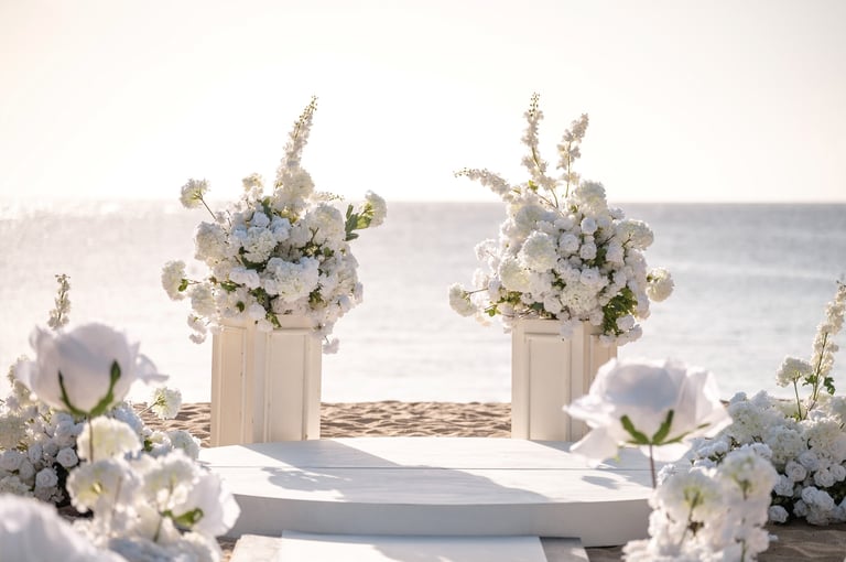 Beach wedding ceremony setup with white floral arrangements in pedestals and large white flowers lining an aisle overlooking the ocean