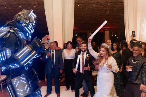 Wedding guests in formal attire dancing with a large blue light-up robot figure at an elegant indoor celebration