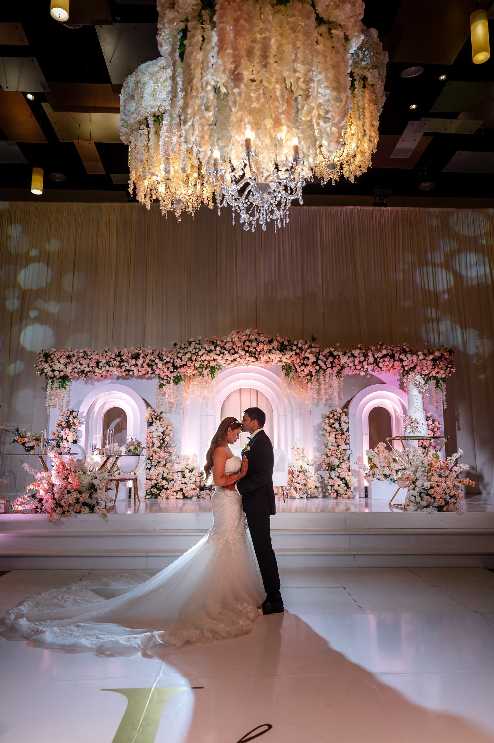 Bride and groom dance together at an elegant indoor wedding reception with ornate floral backdrop and crystal chandelier