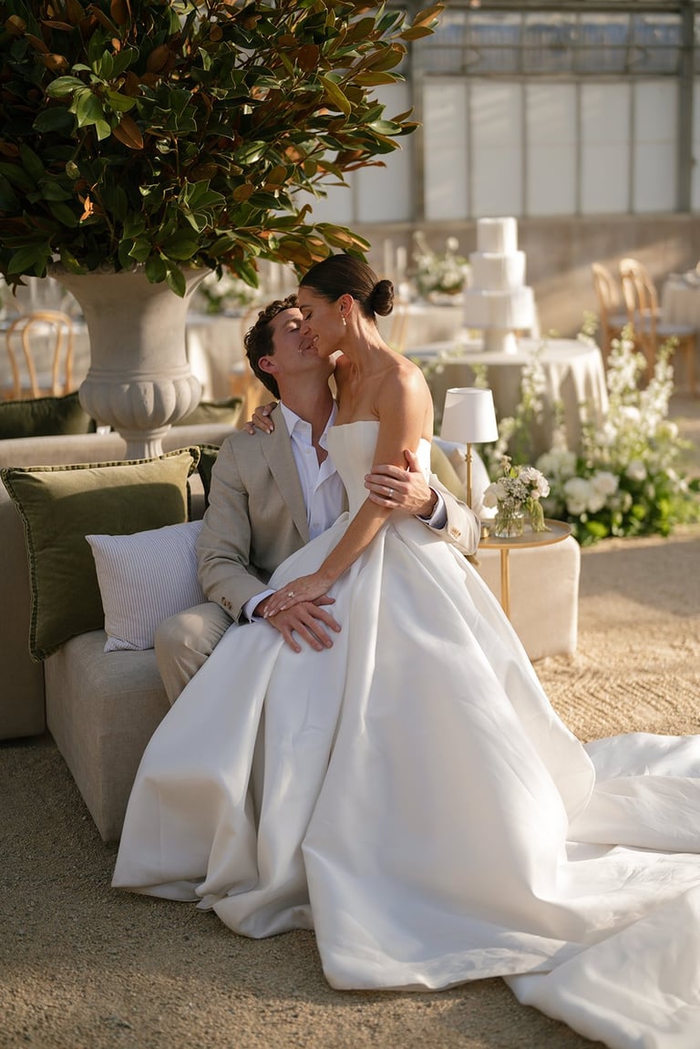 Newlywed couple sitting on a couch, bride in white gown and groom in beige suit, kissing in an elegant decorated room with flowers and natural light