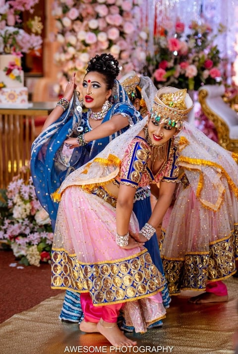 Two women in ornate traditional Indian wedding attire with gold embroidery and jewelry, posing in front of elaborate pink and white floral backdrop