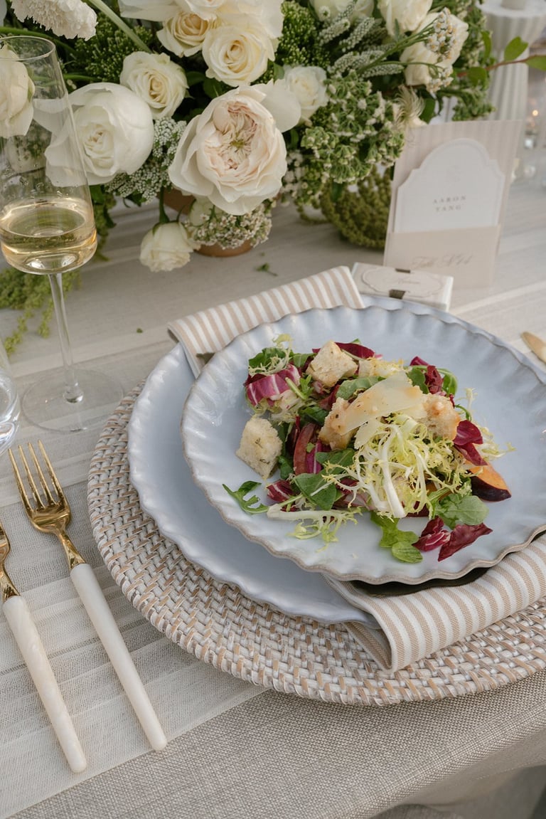 Elegant place setting with a plated salad on blue charger, white roses and greenery centerpiece, wine glass, and gold flatware on linen tablecloth