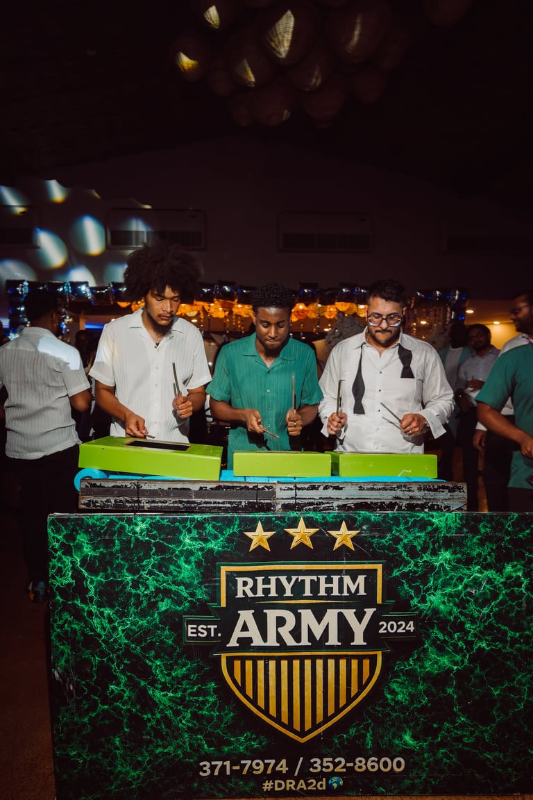 Three men stand behind a DJ booth with Rhythm Army logo at a nighttime event with crowd in background