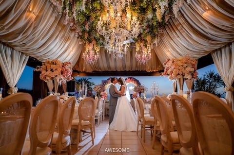 Elegant outdoor wedding ceremony under draped tent with bride and groom, floral arrangements, and gold chairs lining the aisle.
