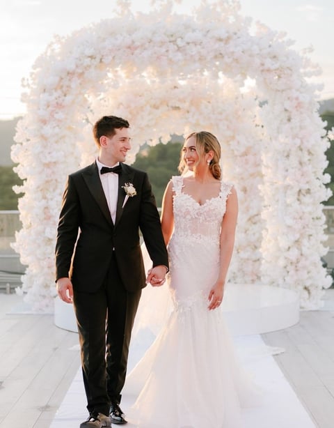 Bride and groom holding hands walking under a large white floral wedding arch on their ceremony day.