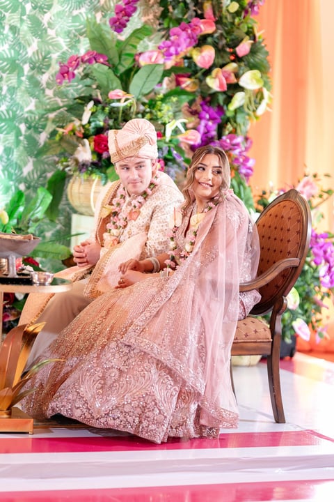 Bride and groom in traditional pink wedding attire sitting on chairs in front of vibrant floral backdrop