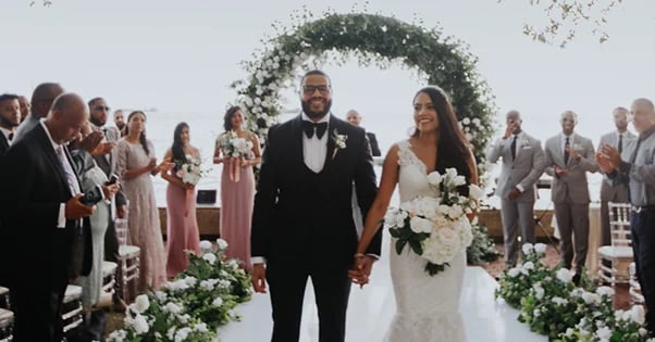 Bride and groom holding hands under a floral arch at their outdoor wedding ceremony with guests watching