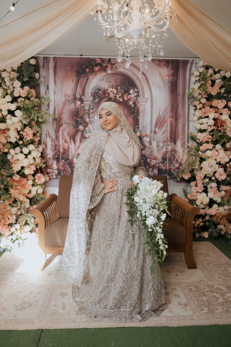 Bride in ornate beaded hijab and gown standing in decorated venue with floral arrangements and chandelier
