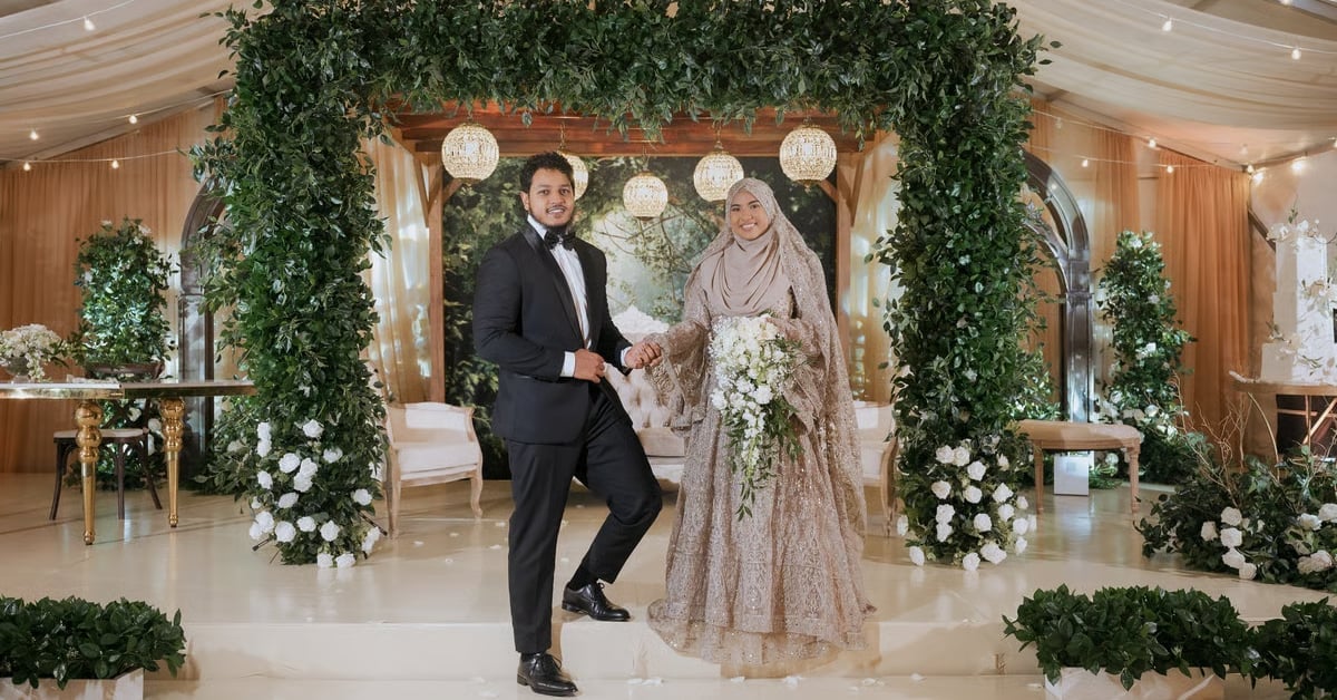 Groom in black tuxedo dancing with bride in beige embellished gown under floral arch with greenery at wedding reception