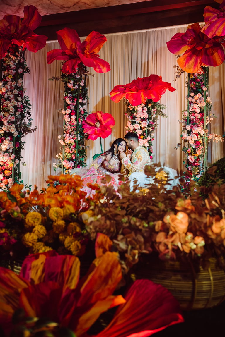 Couple embracing at ornate wedding venue with oversized red and pink floral arrangements and decorative backdrop
