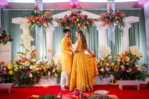 A couple in golden wedding attire stands on a red platform under ornate floral decorations and hanging flower arrangements for a traditional wedding ceremony