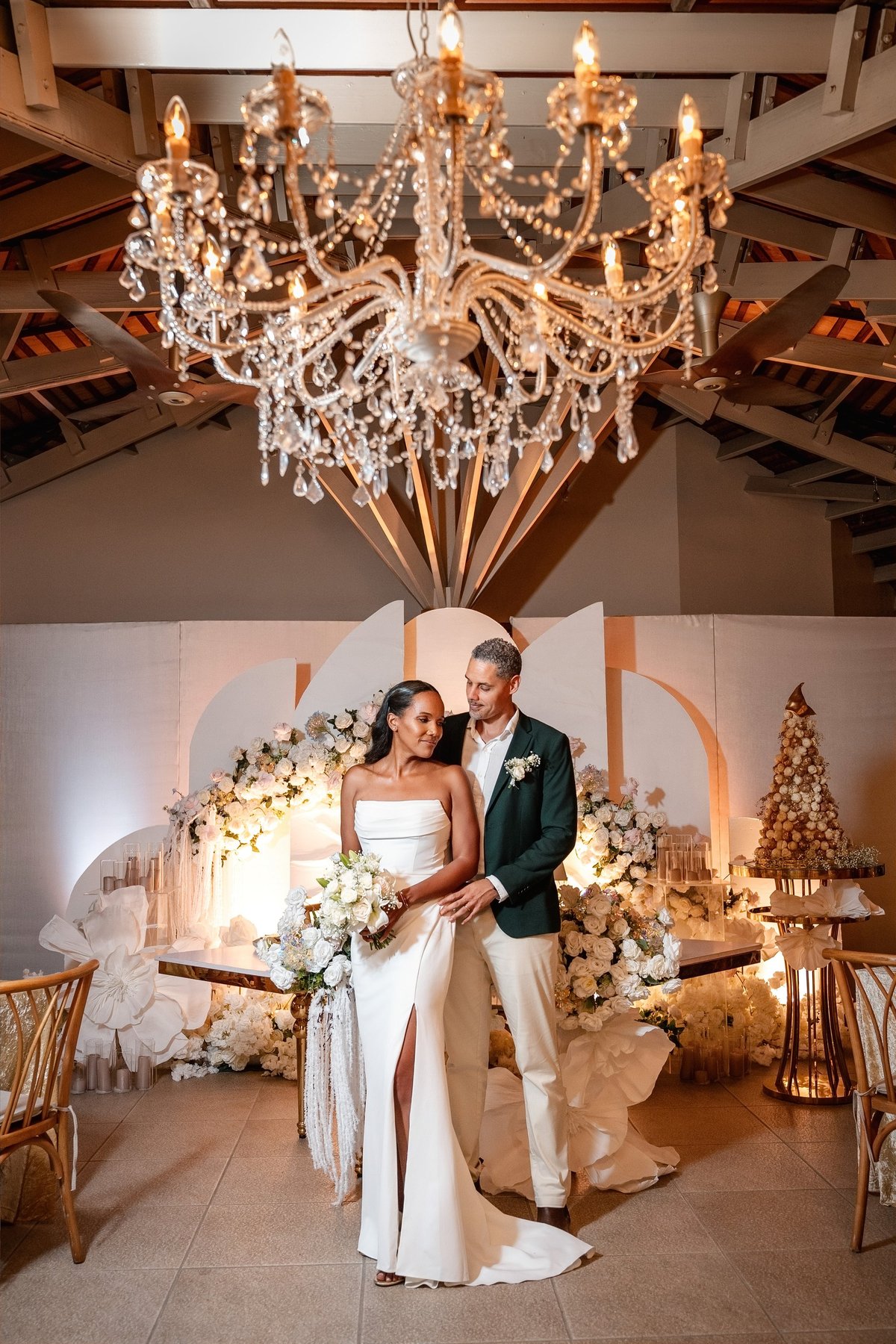 Bride and groom posing in elegant wedding venue under crystal chandelier with floral decorations and gold accents