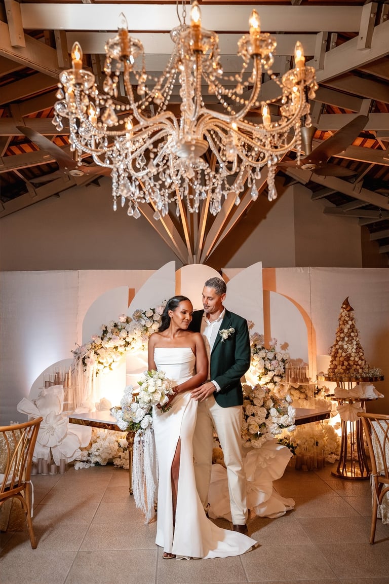Bride and groom posing in elegant wedding venue under crystal chandelier with floral decorations and gold accents