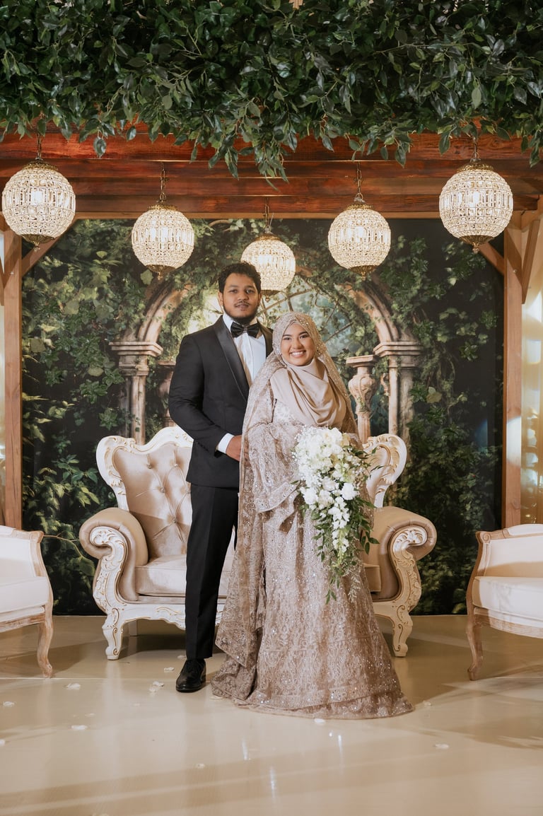 Elegant couple posing at a luxury wedding venue with ornate gold chairs, hanging crystal chandeliers, and decorative green backdrop