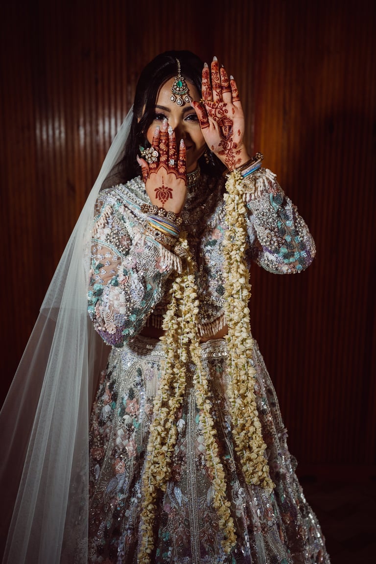 Bride in ornate silver embroidered wedding lehenga with henna-decorated hands, jewelry, and white veil against burgundy backdrop