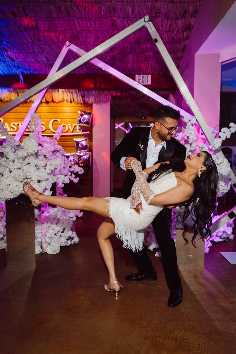 Groom in black suit dips bride in white dress under a white and pink floral-decorated arch at wedding reception