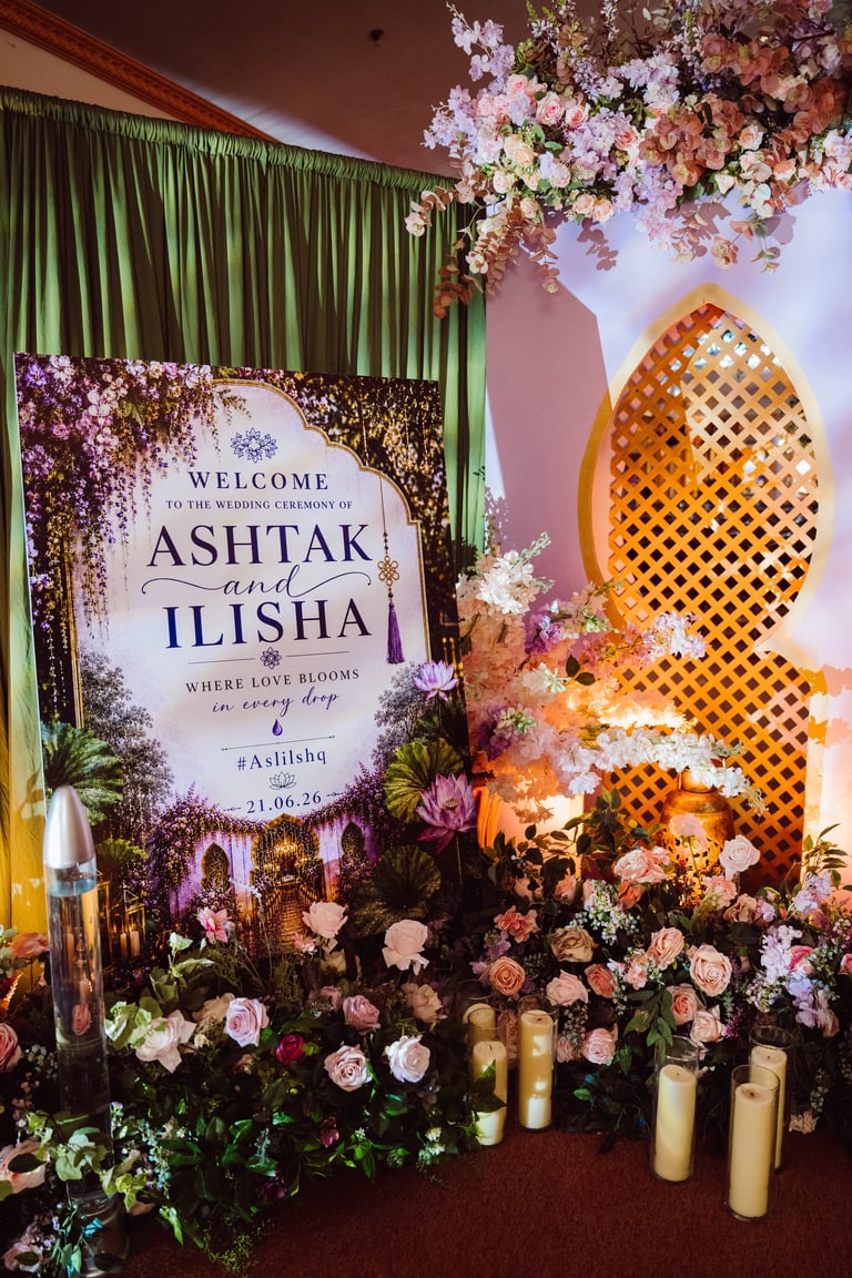 Luxurious wedding entrance decorated with purple and white flowers, a gold leaf arch, welcome sign for Ashtak and Ilisha, and illuminated candles