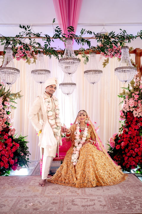 Indian bride and groom in traditional wedding attire posing under ornate crystal chandeliers with floral decorations