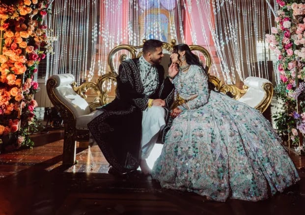 Bride and groom kissing on ornate gold sofa at wedding reception with elaborate floral backdrop and curtains