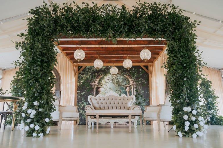 Elegant wedding ceremony setup with beige upholstered chair under wooden archway decorated with cascading greenery, white flowers, and hanging lanterns