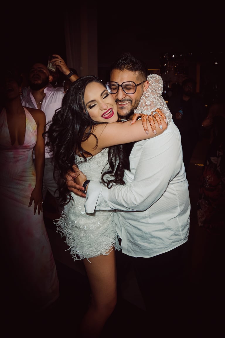 Man and woman embracing at a nighttime party, smiling at the camera with other people visible in the background