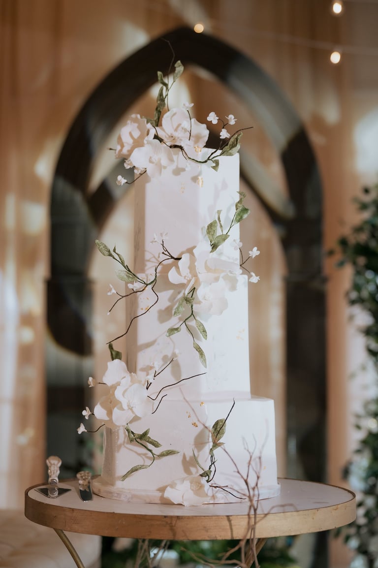 Multi-tiered white wedding cake decorated with pale pink flowers and delicate branches on a wooden table, with an arched backdrop