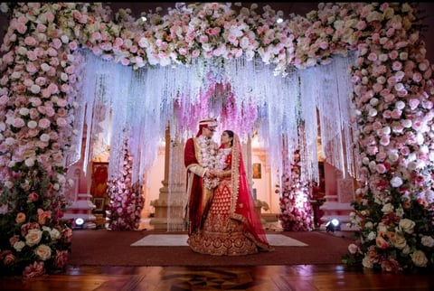 Bride and groom in traditional red wedding attire standing under a lavish floral mandap with pink roses and hanging crystal decorations