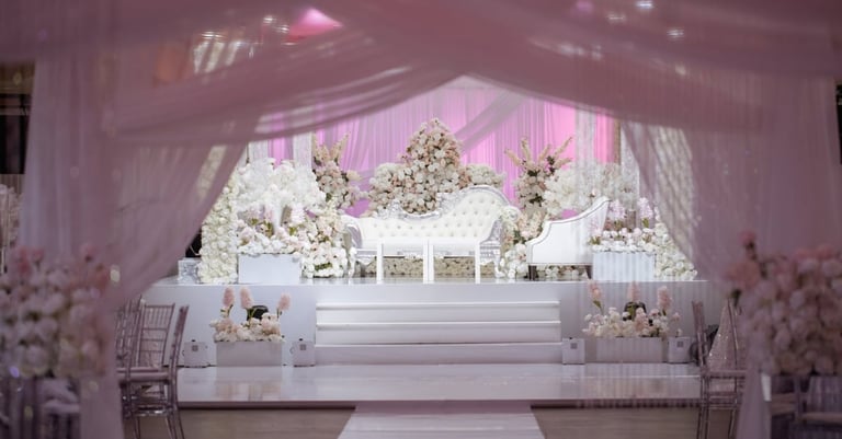 Elegant white wedding stage with pink lighting, white floral arrangements, and sheer drapery